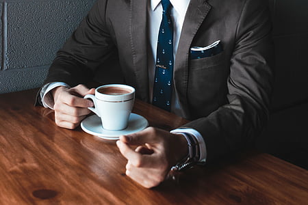 man wearing suit holding a cup of coffee while sitting near table