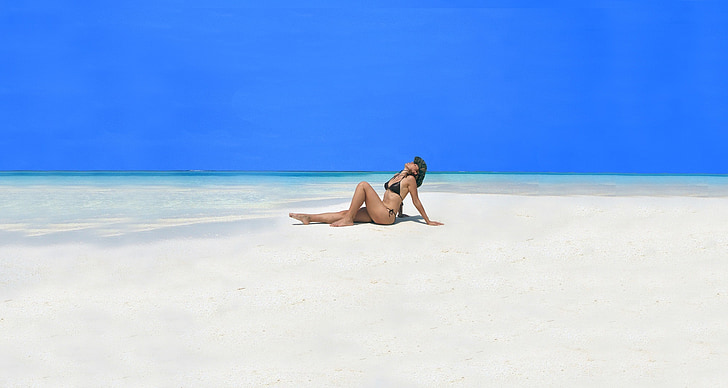 woman wearing black bikini sitting on sand near seashore with hands behind back and looking upwards