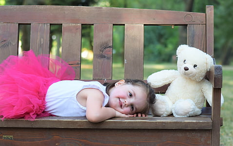 girl lying on brown bench beside bear plush toy at daytime