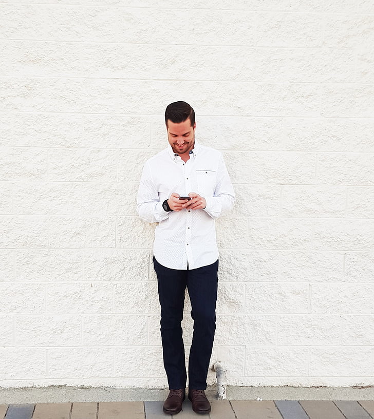 man wearing white dress shirt and black dress pants holding smartphone while leaning to white wall