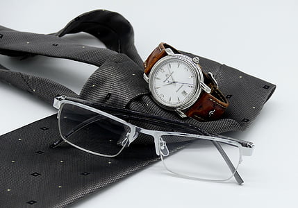 closeup photo of grey framed eyeglasses near round silver-colored analog watch on grey necktie