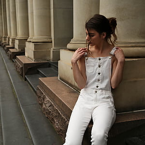 woman wearing white dungaree pants sitting near concrete pillar at daytime