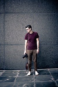 man in maroon crew-neck t-shirt holding Canon DSLR camera standing on the wall