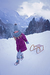 girl pulling sled at her back on snow road near mountains and pine trees covered with snow