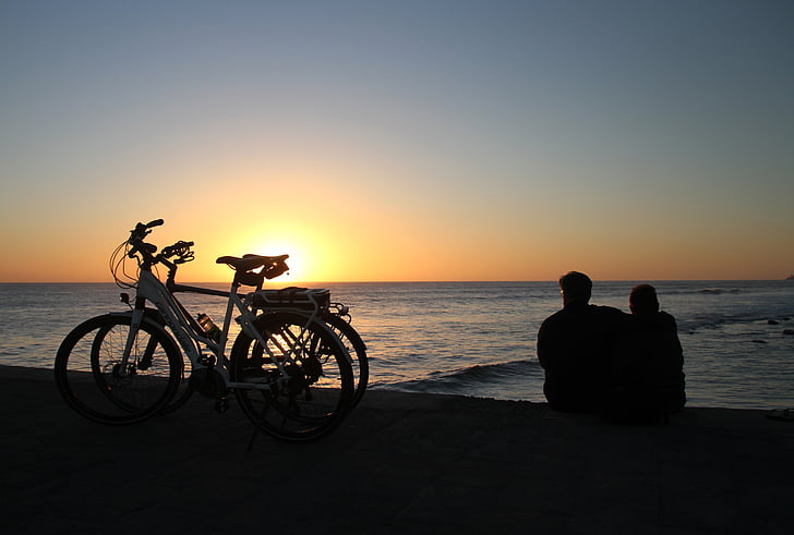 silhouette photography of two person sitting beach shore beside bicycles
