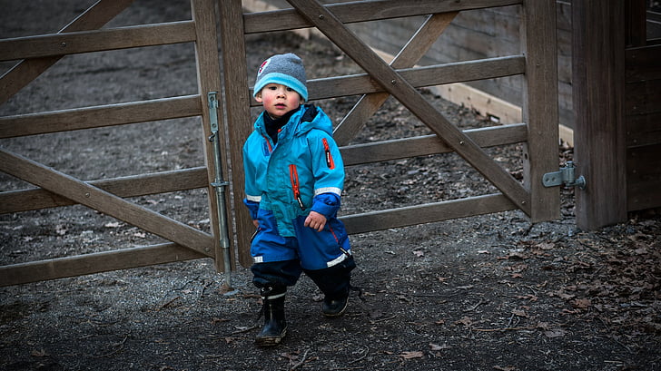 boy wearing blue coat beside brown wooden fence