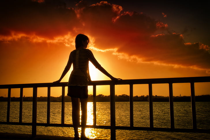silhouette of woman holding on hand rail in front of body of water at golden hour