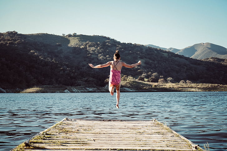 woman jumping on body of water with background of mountains