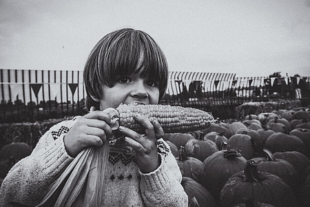 grayscale photo of boy eating corn standing near pumpkins