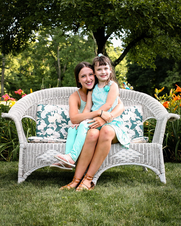 child in teal sleeveless shirt and pants sitting on woman lap in teal sleeveless dress sitting on white wicker sofa