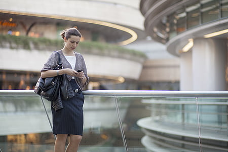 woman in grey 3/4 sleeved blazer and black skirt holding smartphone and black leather tote bag standing