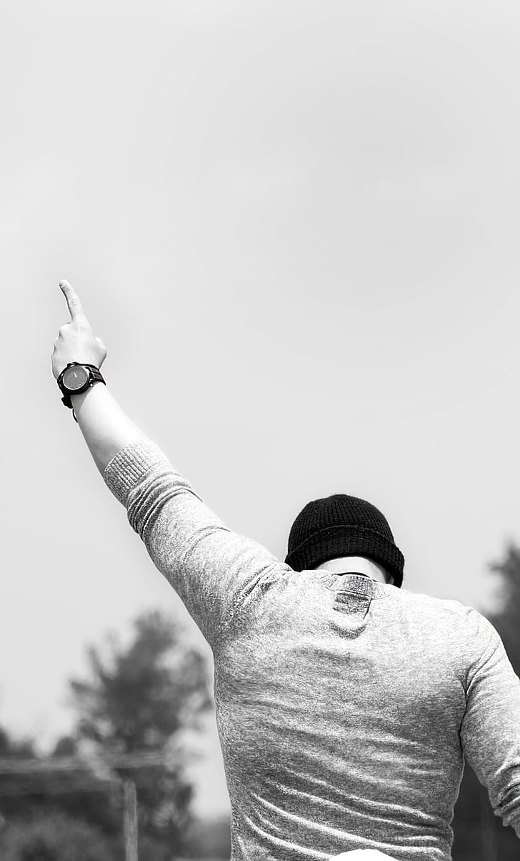 man wearing knit hat pointing his left hand at the sky during day