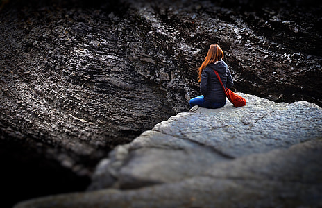 brunette woman wearing black jacket and blue jeans sitting on rocky cliff