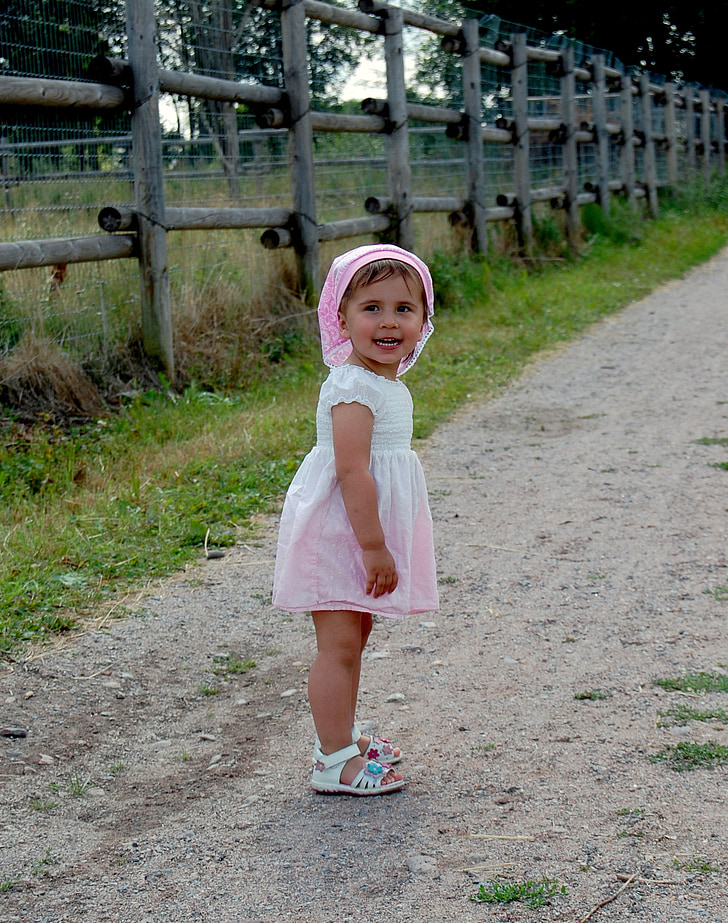 toddler in white and pink dress on brown pathway beside brown fence