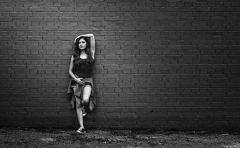 grayscale photo of woman wearing tank top and shorts with jacket on waist leaning on wall