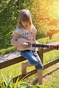girl wearing white t-shirt and blue denim shorts playing brown ukulele leaning on brown wooden fence during daytime