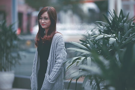 selective focus photo of woman wearing black dress, black framed eyeglasses, and white knit coat beside green plant