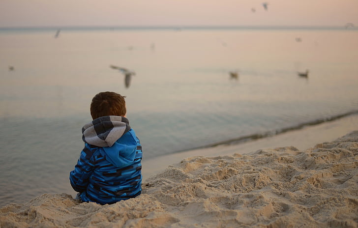 toddler sitting on sand beside body of water during daytime