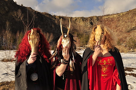 three woman wearing capes holding animal skulls standing in front of mountain