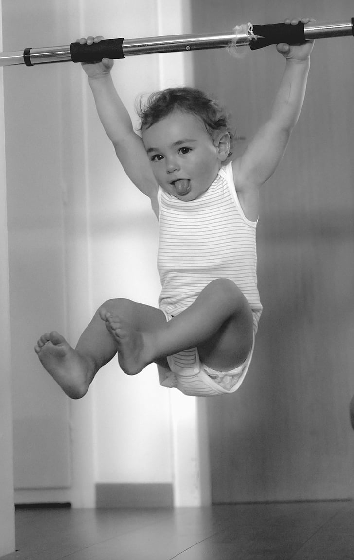 toddler hanging on rod with tongue out grayscale photo