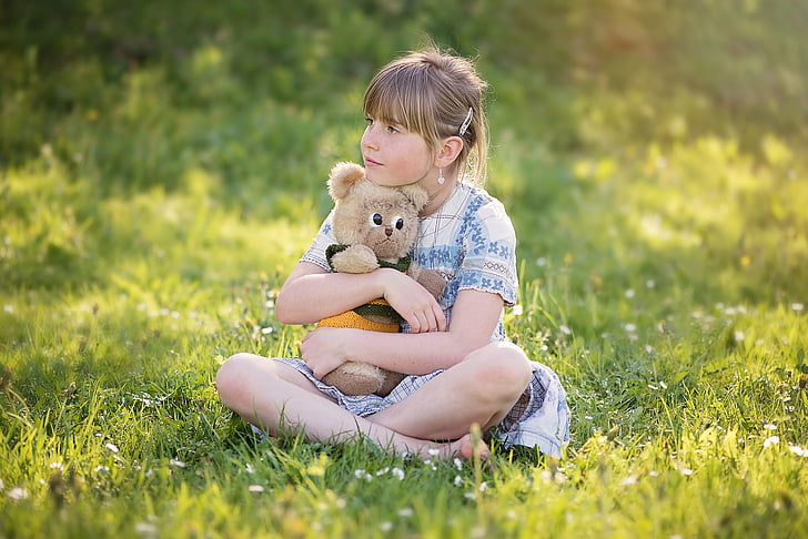 girl hugging brown bear plush toy while sitting on grass field during daytime