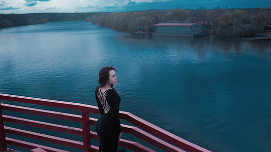 woman in black cage-back dress standing near wooden railings surrounded with body of water