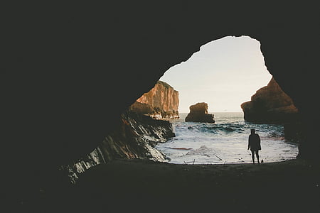 silhouette of person standing under cave near seashore during daytime