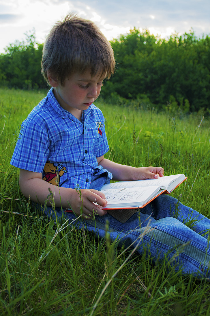 boy sitting on green grass field while reading book during daytime